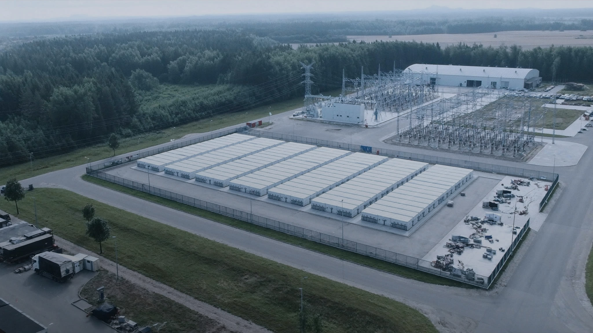 Illustrative view: a utility-scale battery storage yard beside a high-voltage substation, surrounded by forest and farmland in Latvia